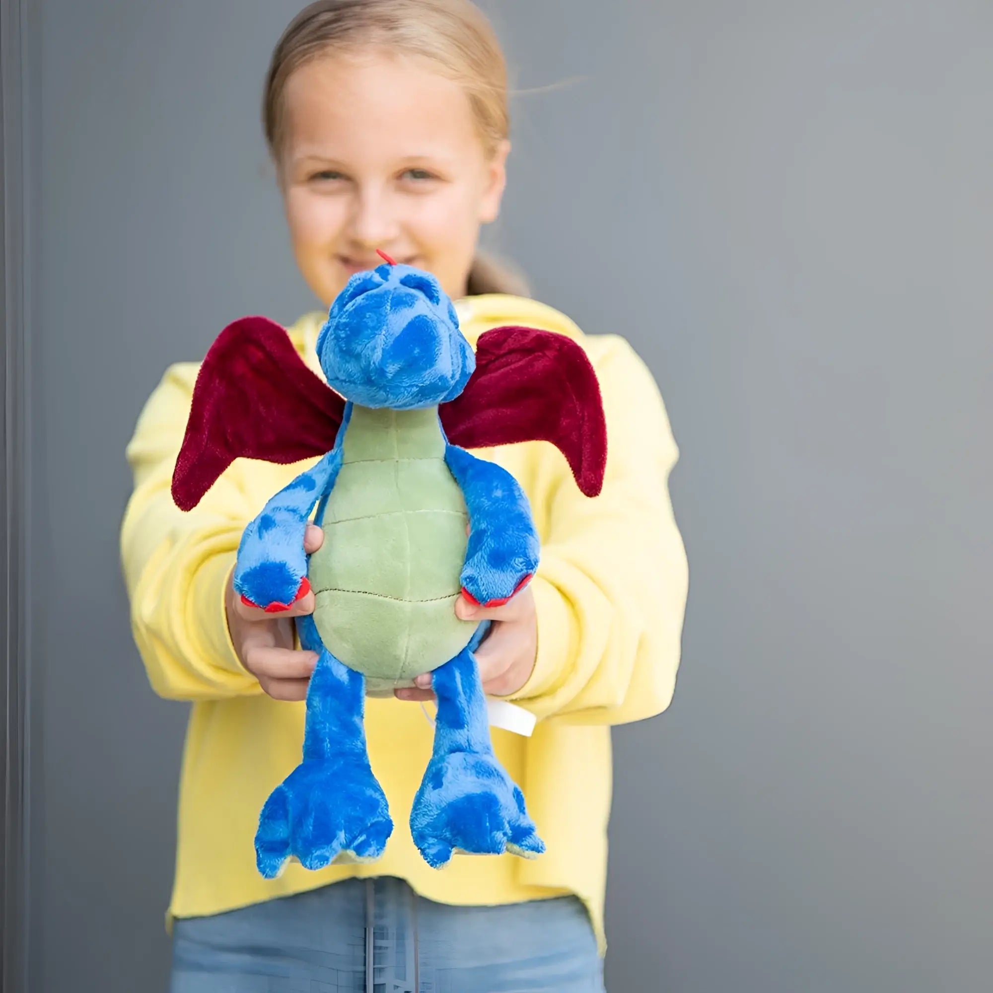 Ein kinderhand haltet den mini feet drache magnus kuschelfreund plüschtier blau mit roten flügeln und grünem bauch