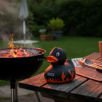 Black rubber duck mit orangeflamme und auftragsgriller branding auf holztisch neben grill im auftragsgriller grillmeister quietsche-ente produkt