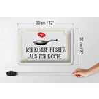 Weißes rechteckiges blechschild mit spruch ich küsse besser als ich koche in schwarz, pfannenillustration und goldrand