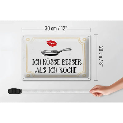 Weißes rechteckiges blechschild mit spruch ich küsse besser als ich koche in schwarz, pfannenillustration und goldrand