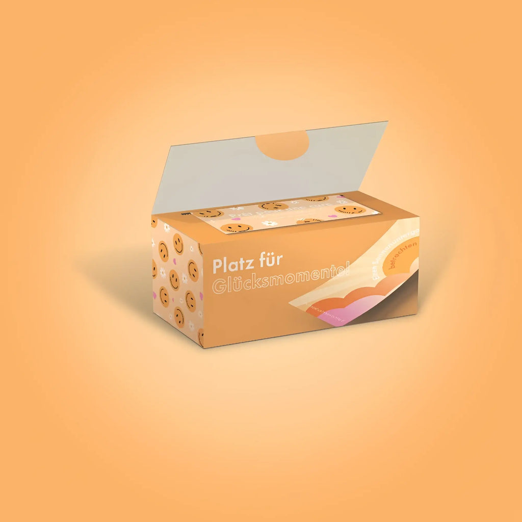 Eine orange kartonbox mit lächelnden keksillustrationen und dem spruch platz für glücksmoment für das kartenspiel challenge-box 30 glücks herausforderungen deutsche version