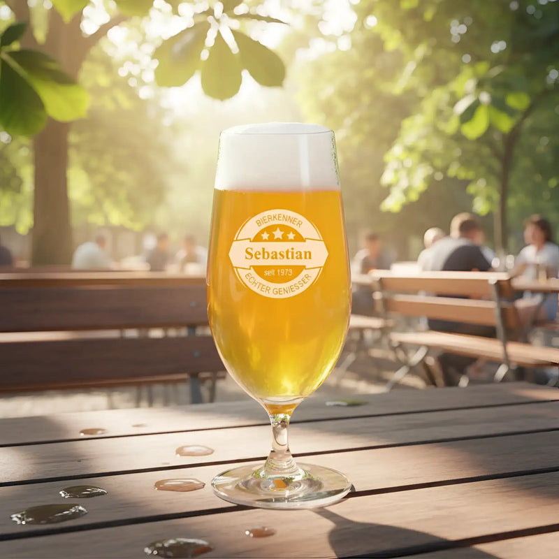 Ein klares, hohes Bierglas mit schaumiger goldener Flüssigkeit und einem eingravierten, personalisierten Etikett, das „sebastian seit 1973“ liest, steht draußen auf einem Holztisch.