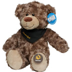 Ein brauner plüschiger oktoberfest bär bodo mit schwarzem bandana und goldener schrift sitzt aufrecht mit beigen pfotenflächen