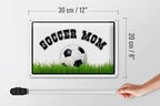 Weißes rechteckiges blechschild 20x30cm mit schwarzem soccer mom text und fußball auf grünem gras